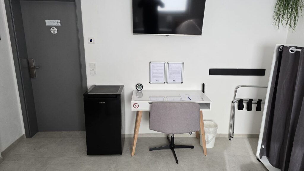 Zimmer mit Schreibtisch, Stuhl, Fernseher, Kühlschrank und Gepäckablage